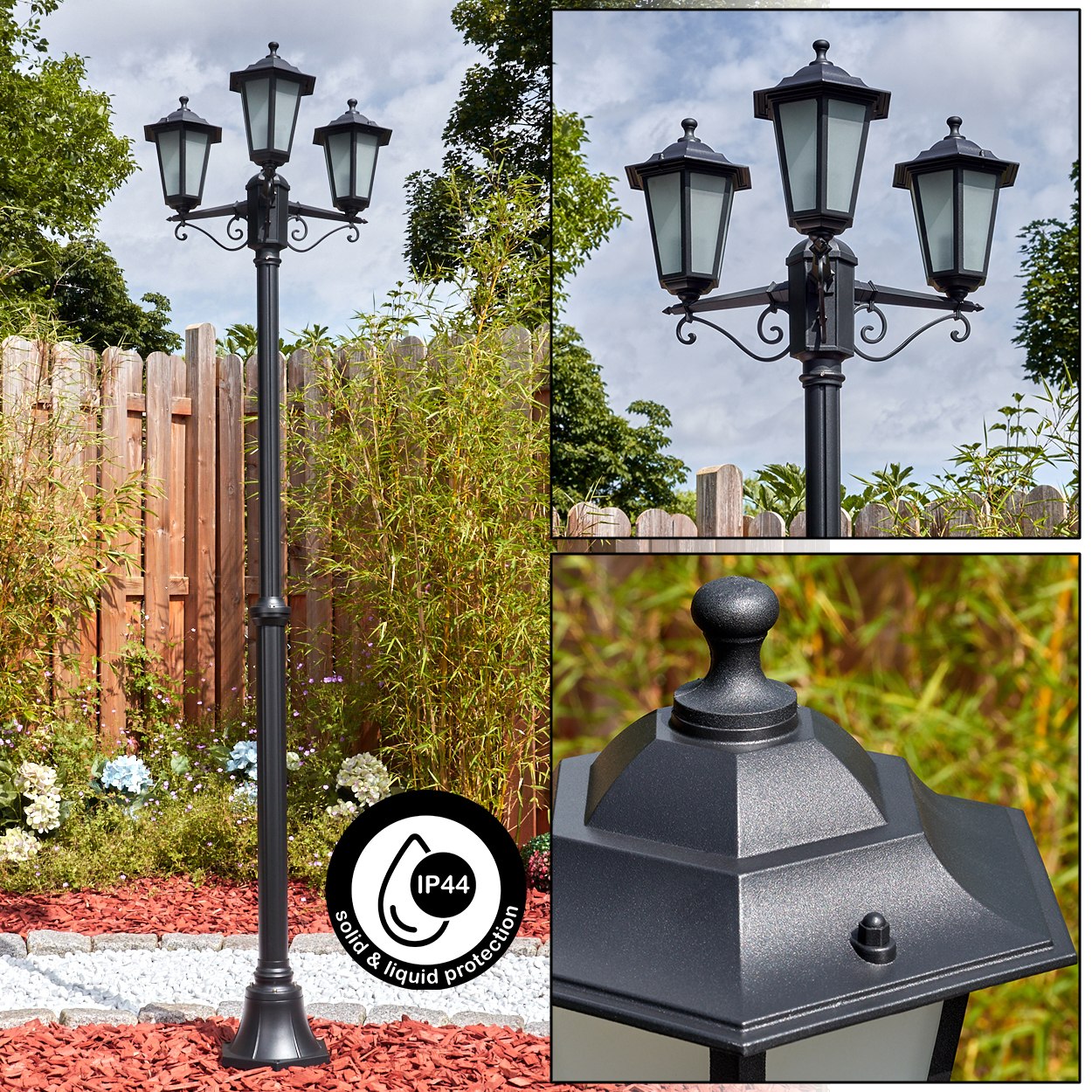 Eine schwarze Außenlampe mit drei Lichtern. Sie befindet sich in einem Garten mit grünen Pflanzen, einem Holzzaun und roter Mulche.