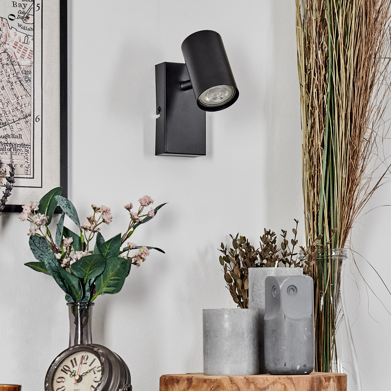 Eine schwarze Wandleuchte an einer weißen Wand. Darunter Blumen in einer Vase, eine Uhr und dekorative Gegenstände.