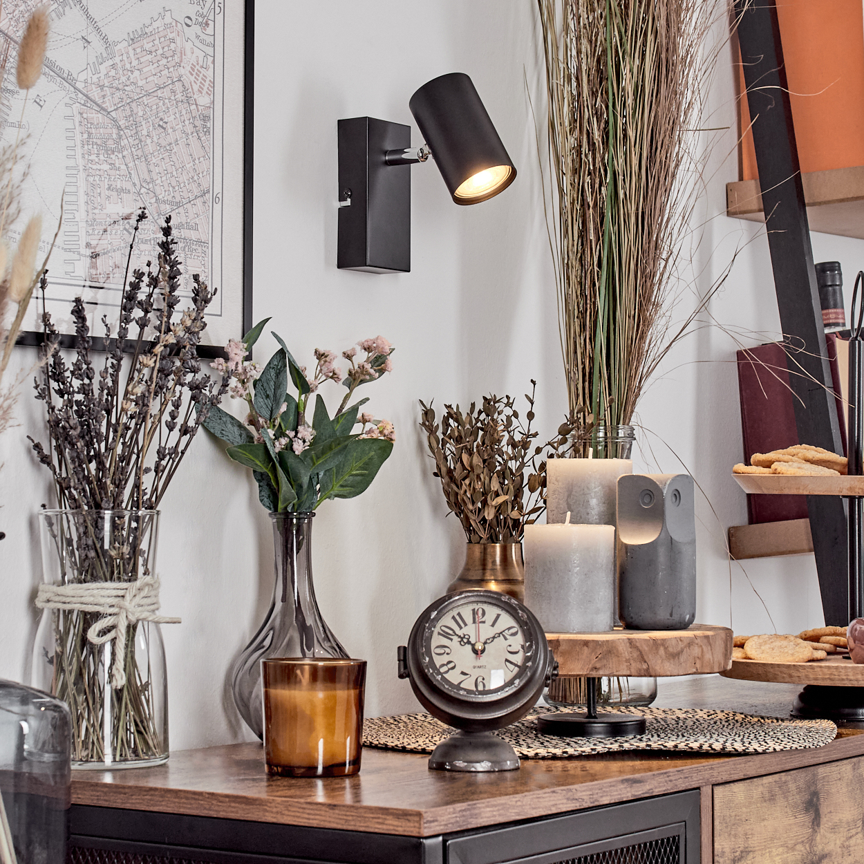 Raumecke mit Deko: eine Karte, eine Lampe, Blumen in Vasen, eine Uhr, Kerzen und Kekse auf einem Holzregal.