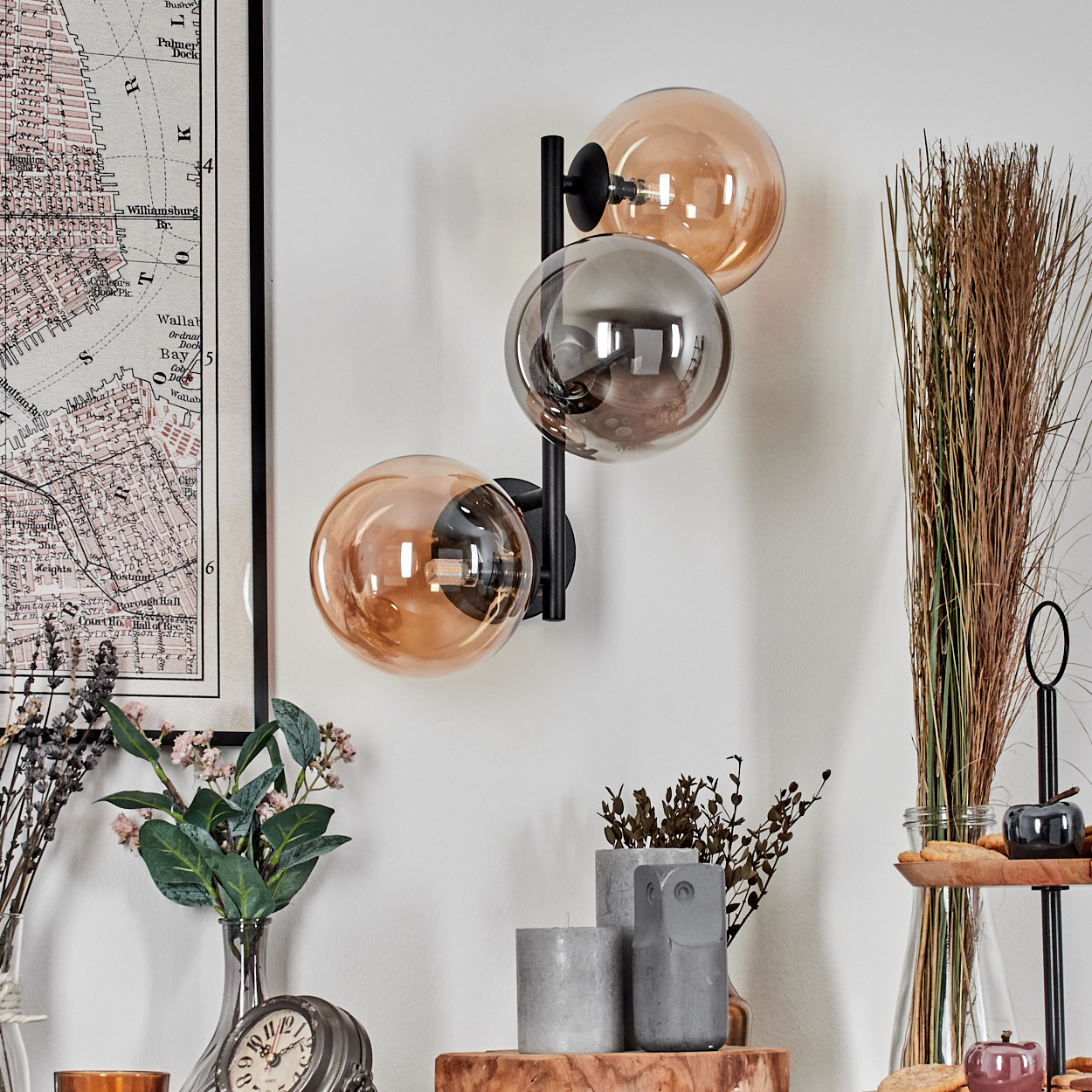 An einer weißen Wand ist eine Wandlampe mit drei Glaskugeln angebracht. Weitere Dekorationen sind eine Karte, Blumen, Kerzen und Dekorationsartikel.