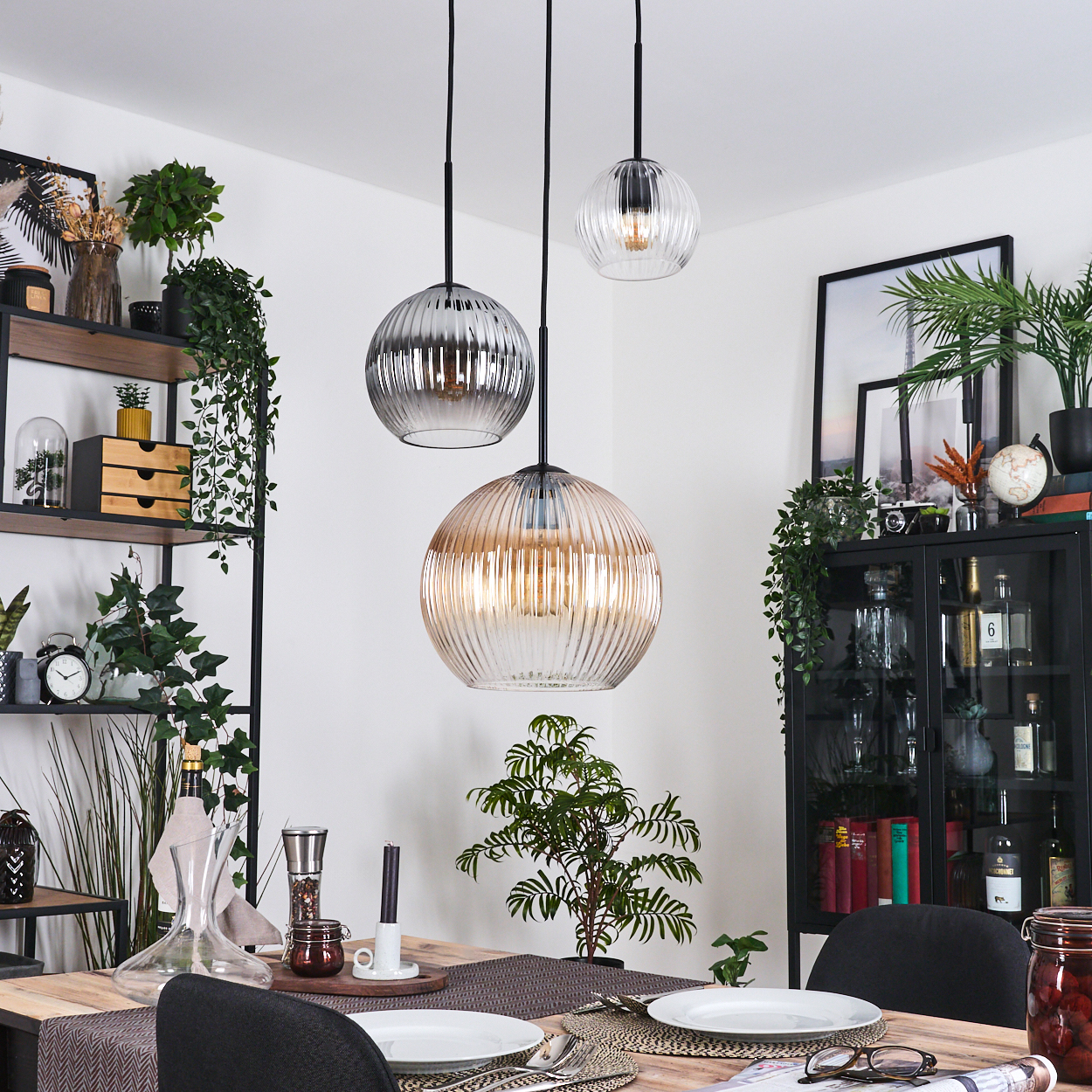 Esszimmer mit drei Hängelampen, Tisch mit Tellern, Pflanzen und einem schwarzen Schrank mit verschiedenen Gegenständen.