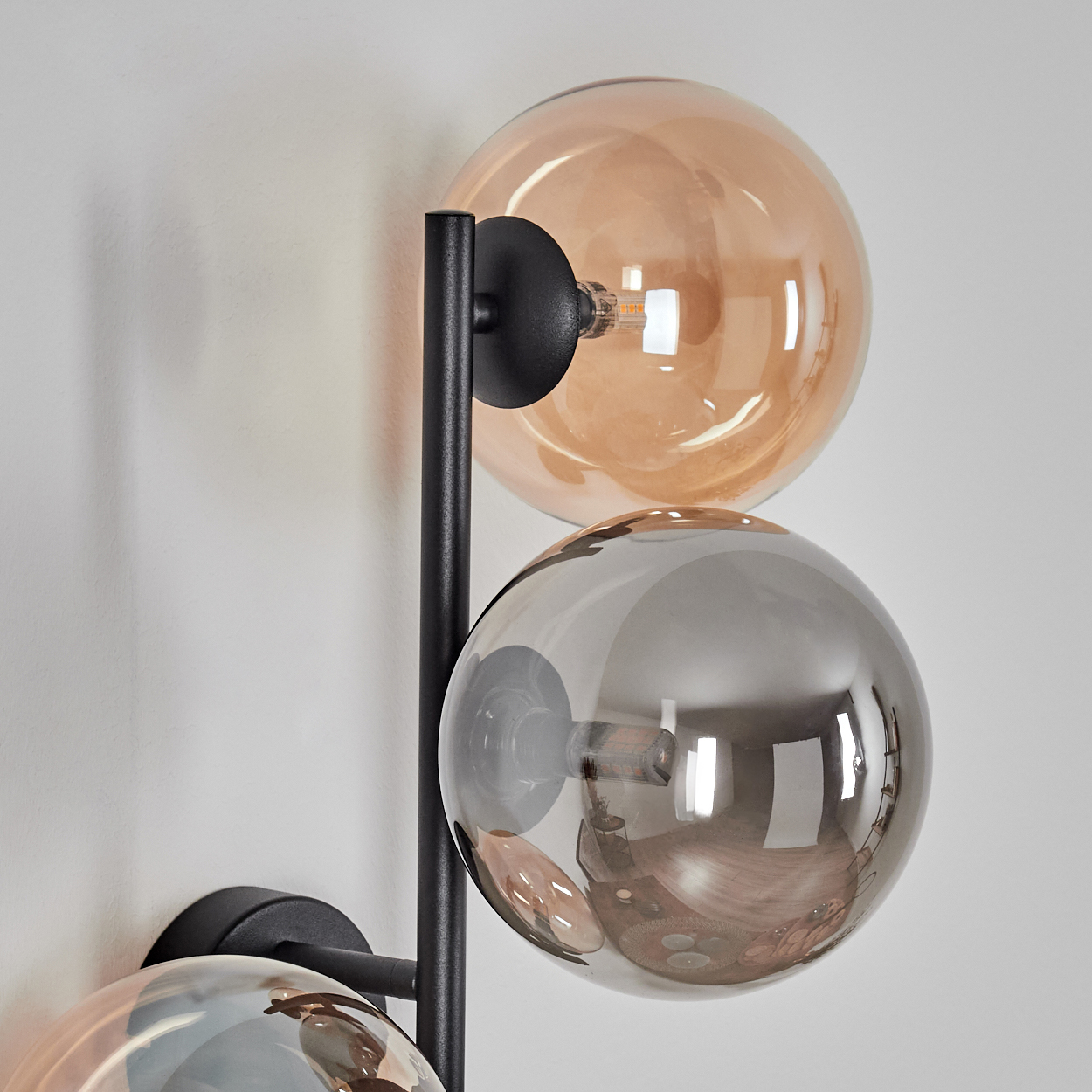 Eine moderne Wandlampe mit drei Glaskugeln in Bernstein und Grau. Die Lampe hat ein schwarzes Metallgestell, das an einer weißen Wand befestigt ist.