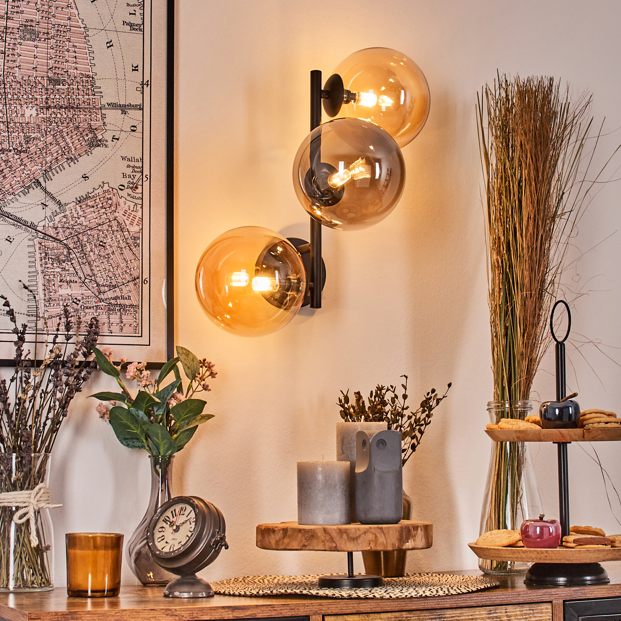 Eine Wandlampe mit drei Glaskugeln. Auf einem Holztisch mit Kerzen, einer Uhr und Keksen befinden sich eine Karte und Vasen mit Blumen.