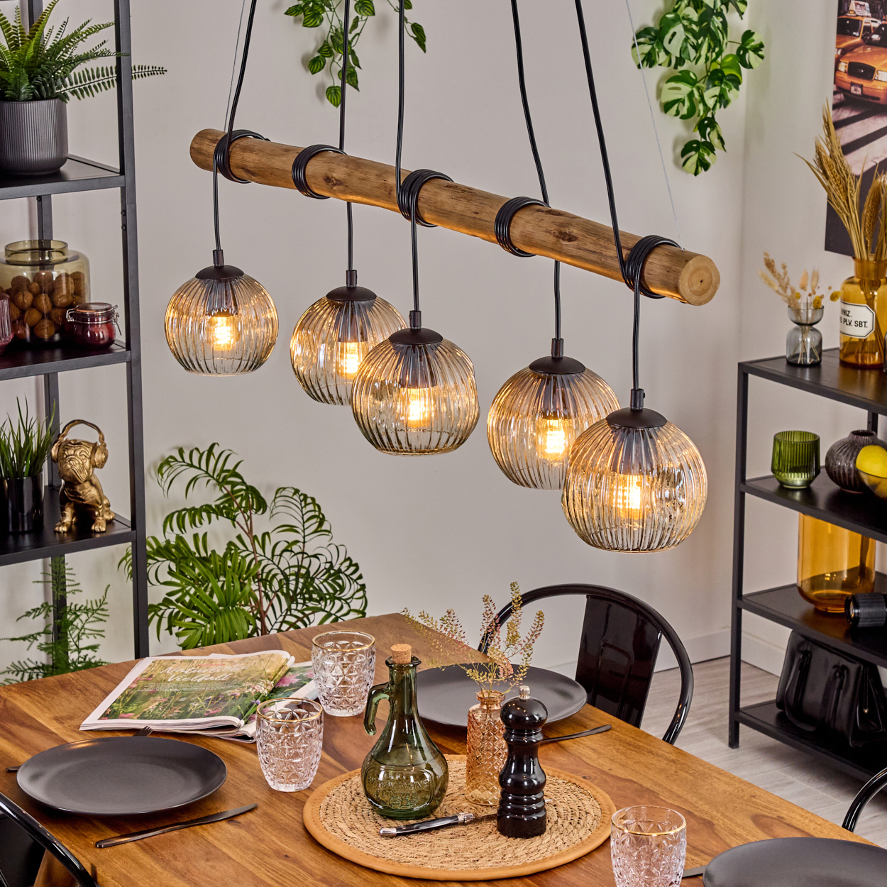 Esszimmer mit Holztisch, Hängelampen und Dekoration. Tisch gedeckt mit Tellern, Gläsern und einer Flasche. Regale mit Pflanzen.
