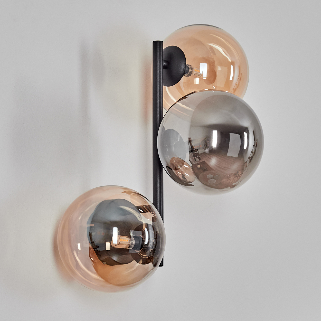 Eine moderne Wandlampe mit drei kugelförmigen Schirmen. Die Schirme sind bernsteinfarben und silber. Die Lampe ist an einem schwarzen Metallrahmen befestigt.