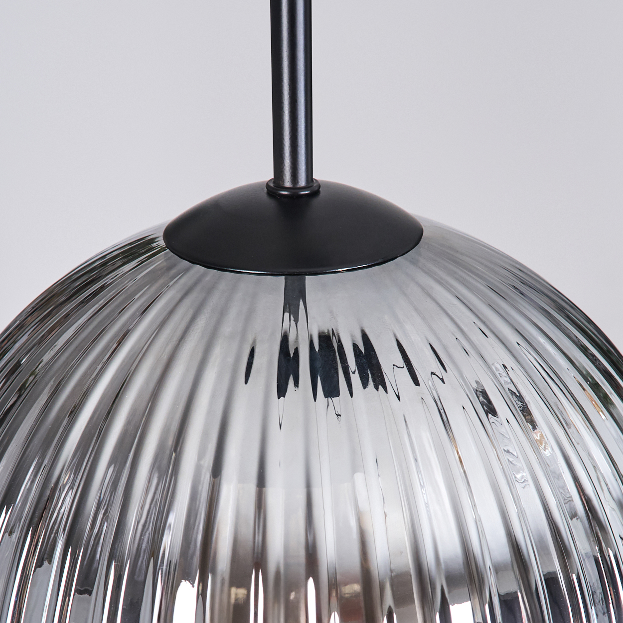 Nahaufnahme eines Glasschirms mit vertikalen Rillen, der Licht reflektiert. Er hängt an einer schwarzen Metallhalterung vor weißem Hintergrund.