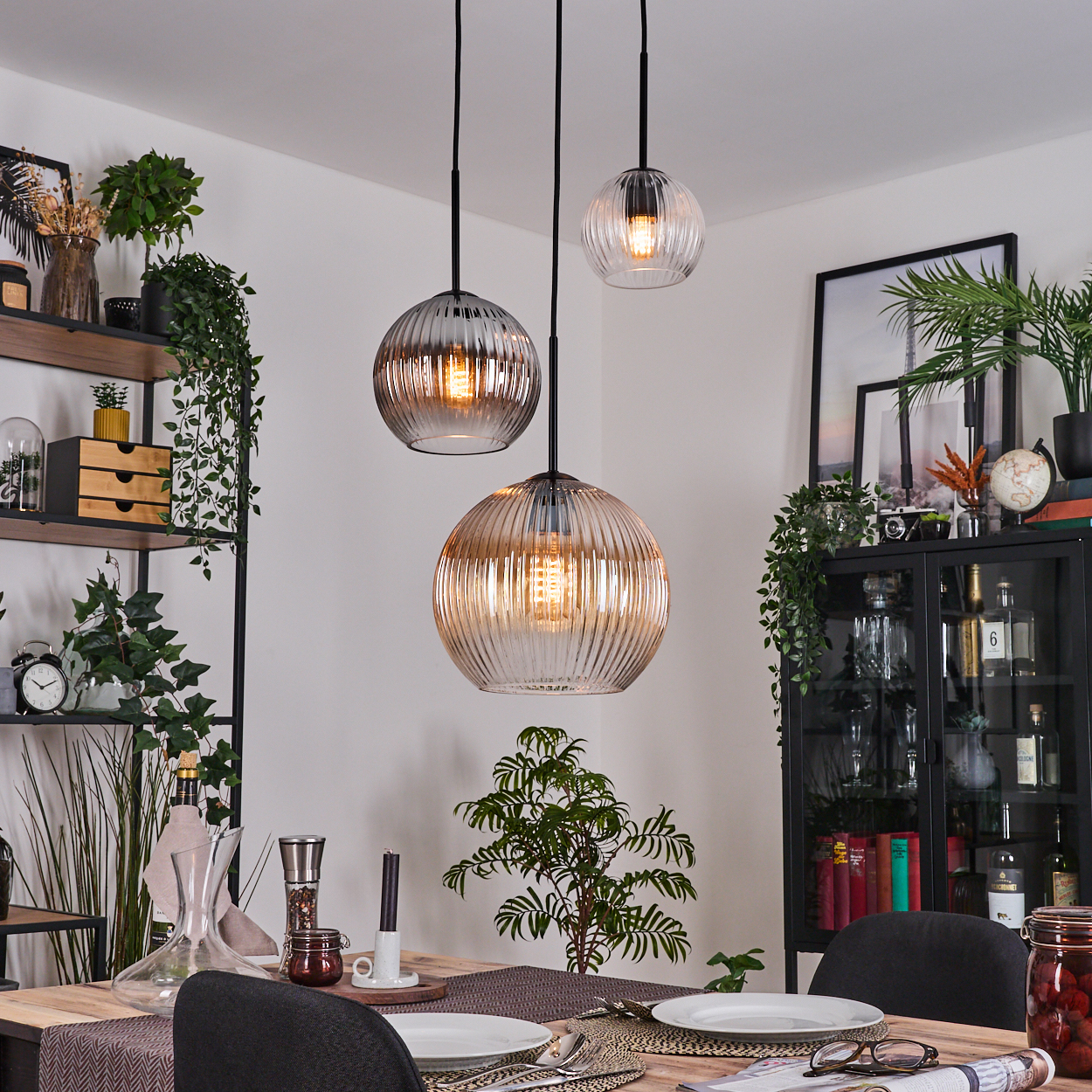 Esszimmer mit drei Hängelampen. Tisch mit Tellern, Gläsern. Pflanzen auf Regalen und Schränken. Schwarze Stühle.
