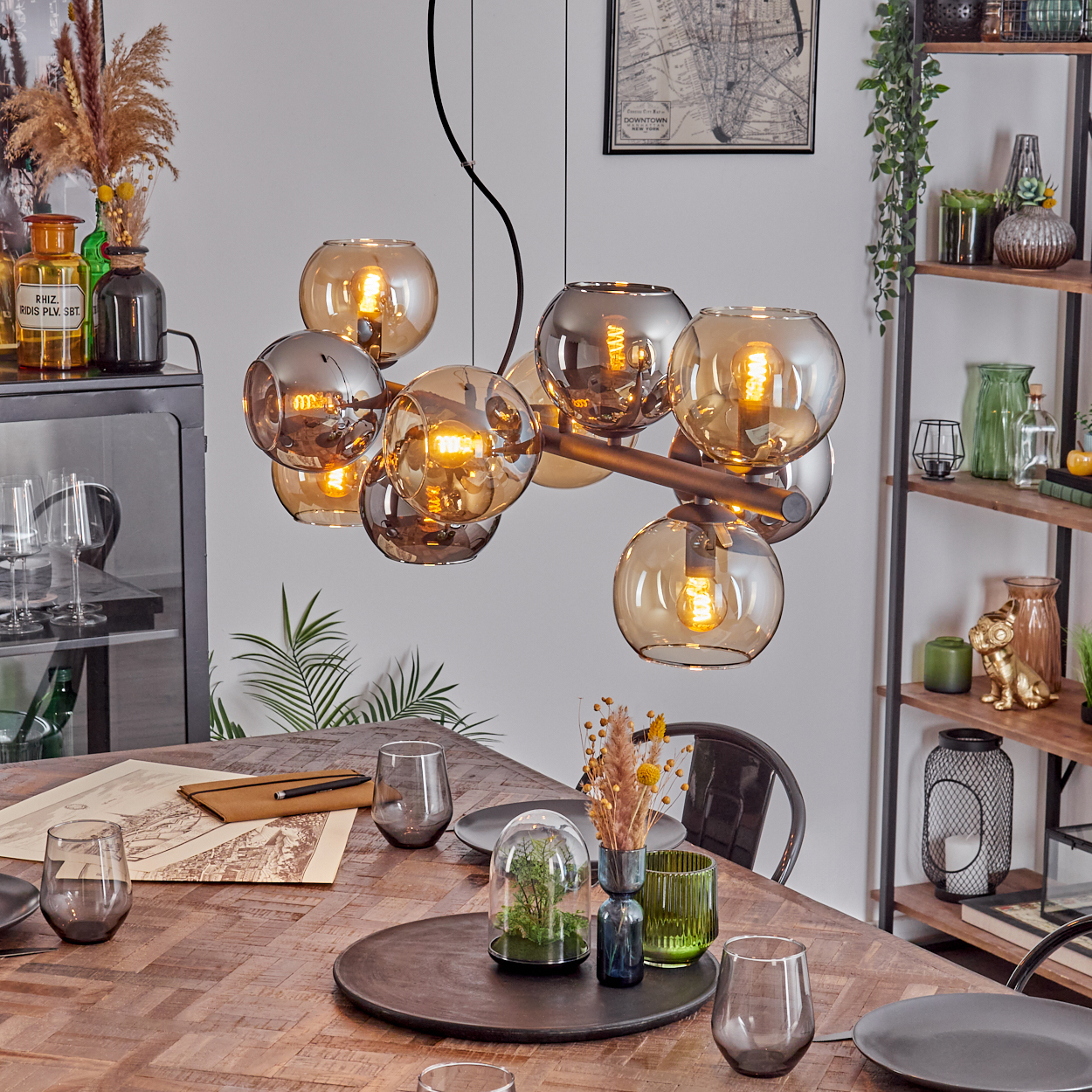 Esszimmer mit Holztisch, modernem Kronleuchter und Dekoartikeln. Teller, Gläser und ein Terrarium stehen auf dem Tisch.