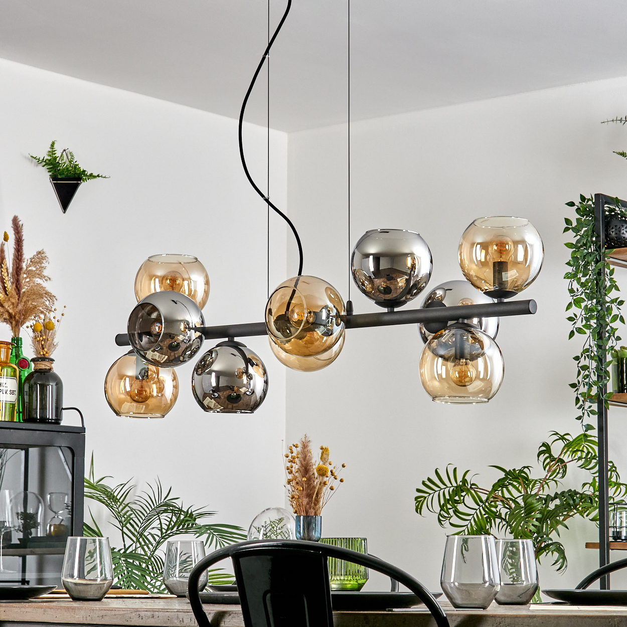 Ein modernes Esszimmer mit einem schwarzen Kronleuchter mit Glaskugeln, Pflanzen und einem Tisch mit Gläsern.