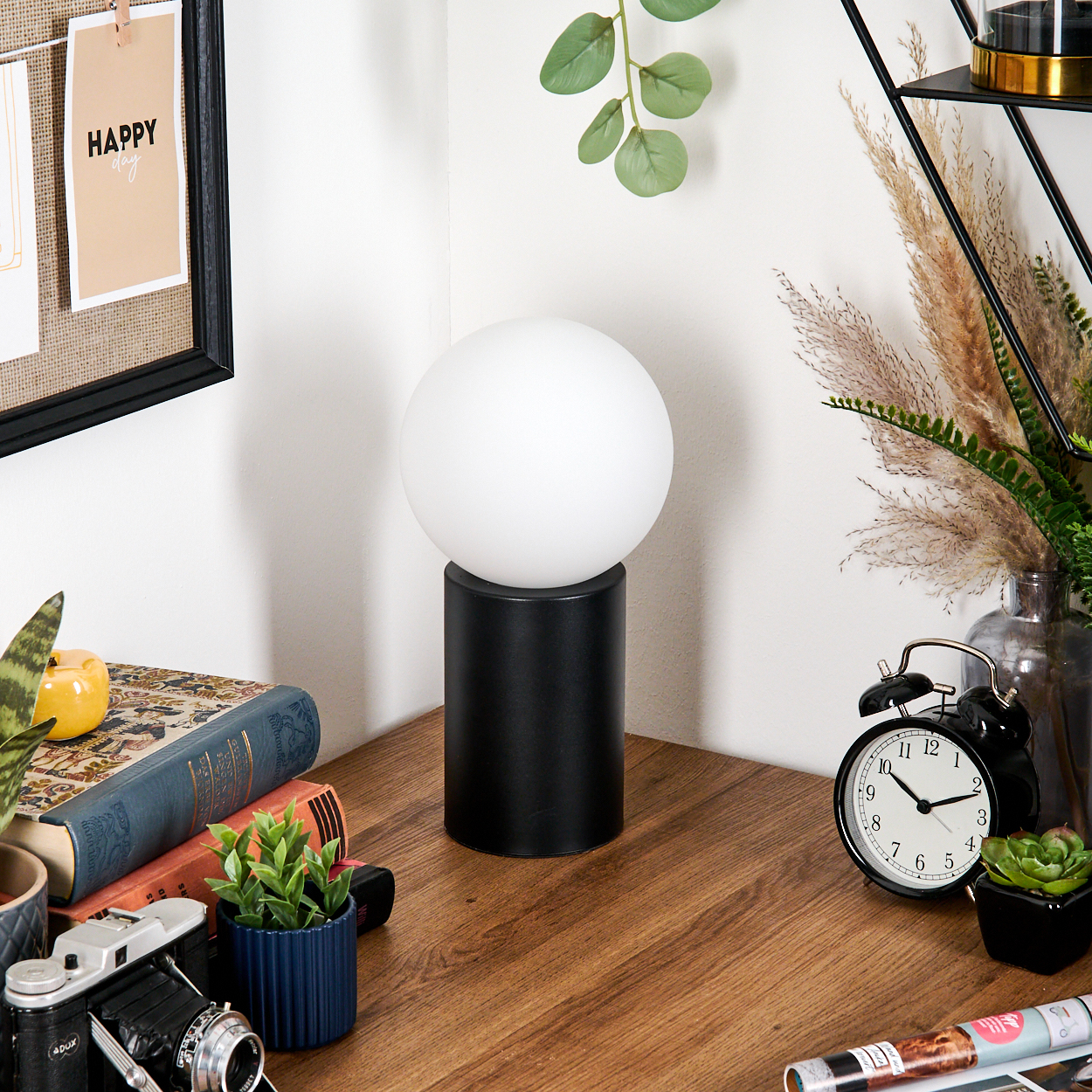 Ein Schreibtisch mit Lampe, Büchern, Kamera, Pflanzen und Wecker. Ein 'Happy Day'-Zettel ist an der Wand befestigt. Die Szene ist hell.