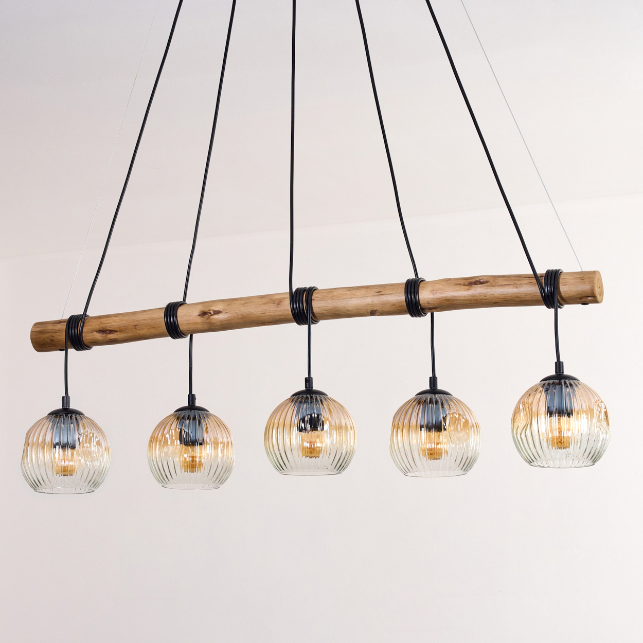 Eine moderne Pendelleuchte mit fünf Glaskugelleuchten, die von einem Holzbalken abgehängt sind. Der Hintergrund ist eine weiße Wand.