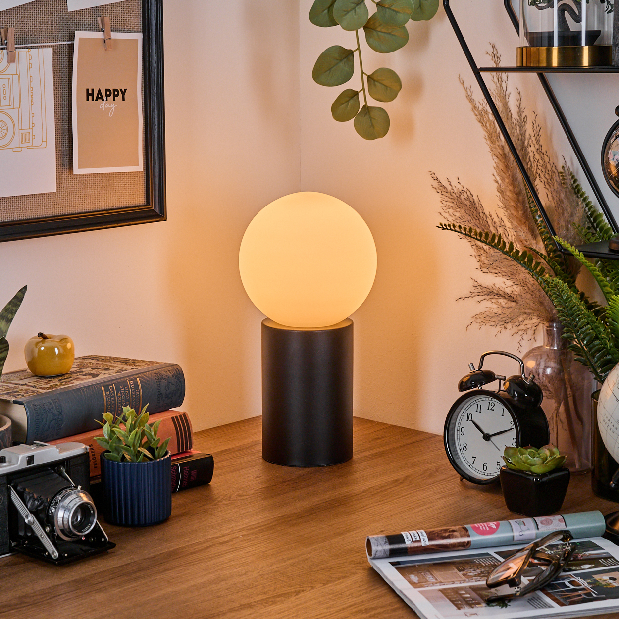 Ein Schreibtisch mit einer leuchtenden runden Lampe, Büchern, einer Kamera, einem Wecker und einer Pinnwand mit Happy Day.