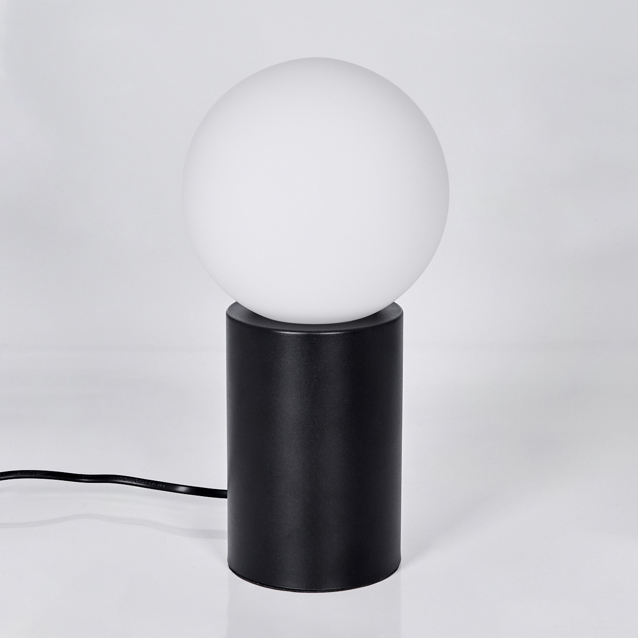 Eine moderne Lampe mit einem weißen, kugelförmigen Schirm auf einem schwarzen, zylindrischen Sockel. Ein schwarzes Netzkabel geht ab.