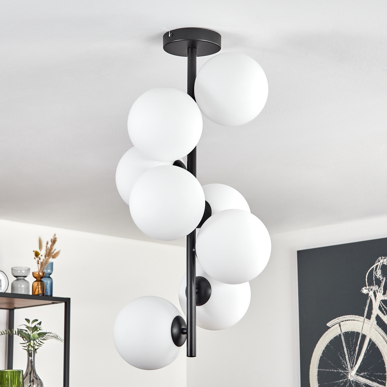 Eine moderne Deckenlampe mit weißen Kugelleuchten. Die Lampe hat einen schwarzen Metallrahmen. Im Hintergrund befindet sich ein Fahrradkunstwerk.