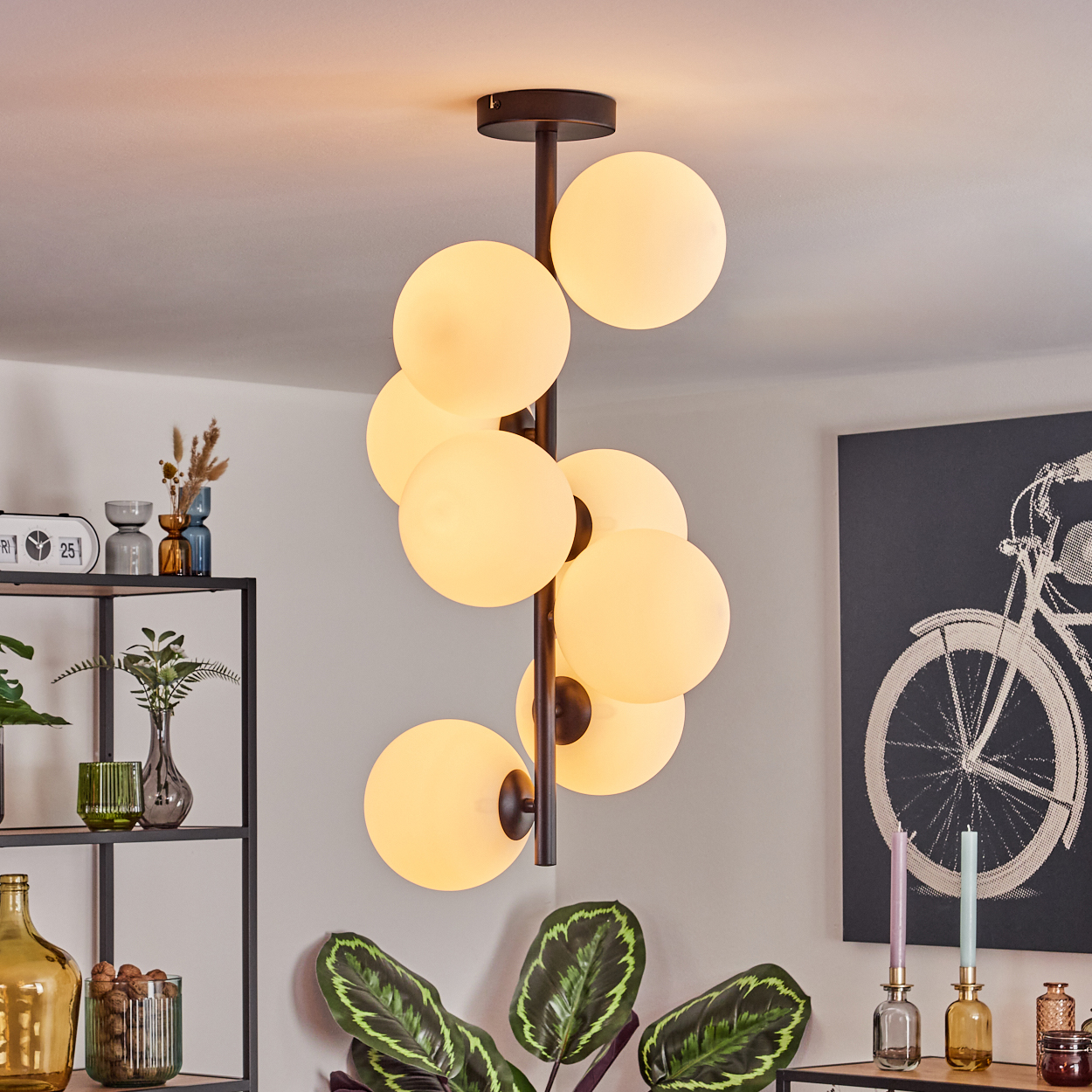 Eine moderne Deckenlampe mit mehreren leuchtenden Kugeln. Rechts ein Gemälde eines Fahrrads. Pflanzen, Vasen und Regale sind ebenfalls sichtbar.