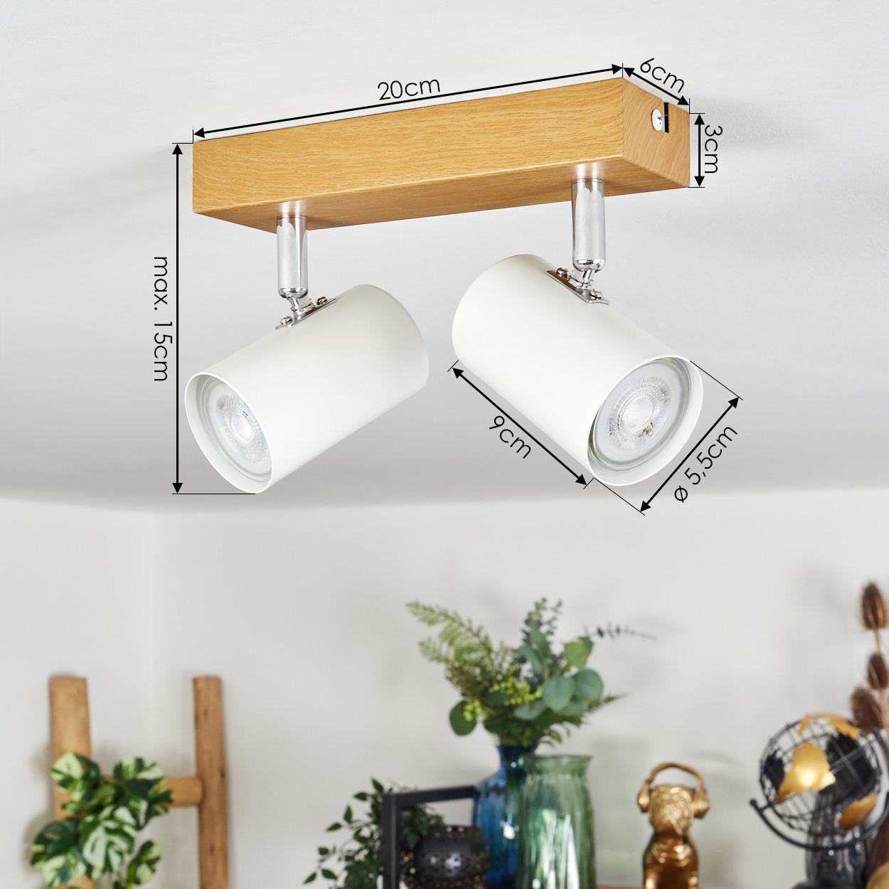 Ein weißer Deckenstrahler mit zwei Lampen. Er hat eine Holzbasis und ist an einer weißen Decke montiert. Abmessungen sind angegeben.