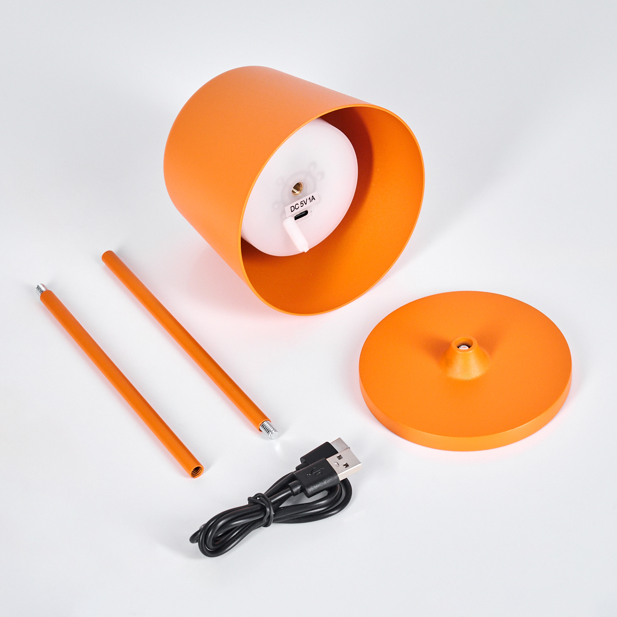 Ein orangefarbenes Lampen-Set mit Teilen auf einer weißen Oberfläche. Enthält Sockel, Zylinder, zwei Stäbe und ein Ladekabel.