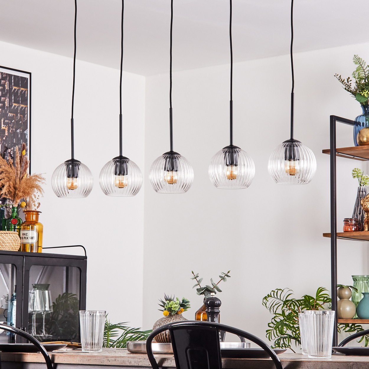 Esszimmer. Fünf Glas-Pendelleuchten hängen über einem Tisch. Es gibt Gläser, Pflanzen und ein schwarzes Metallregal.