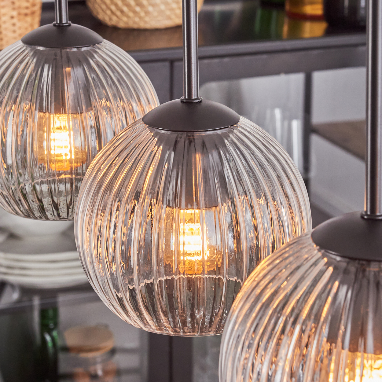 Drei Glas-Kugel-Pendelleuchten mit sichtbaren Glühbirnen. Gerippte Glasschirme, dunkle Metallarmaturen. Hintergrund enthält Küchenartikel.