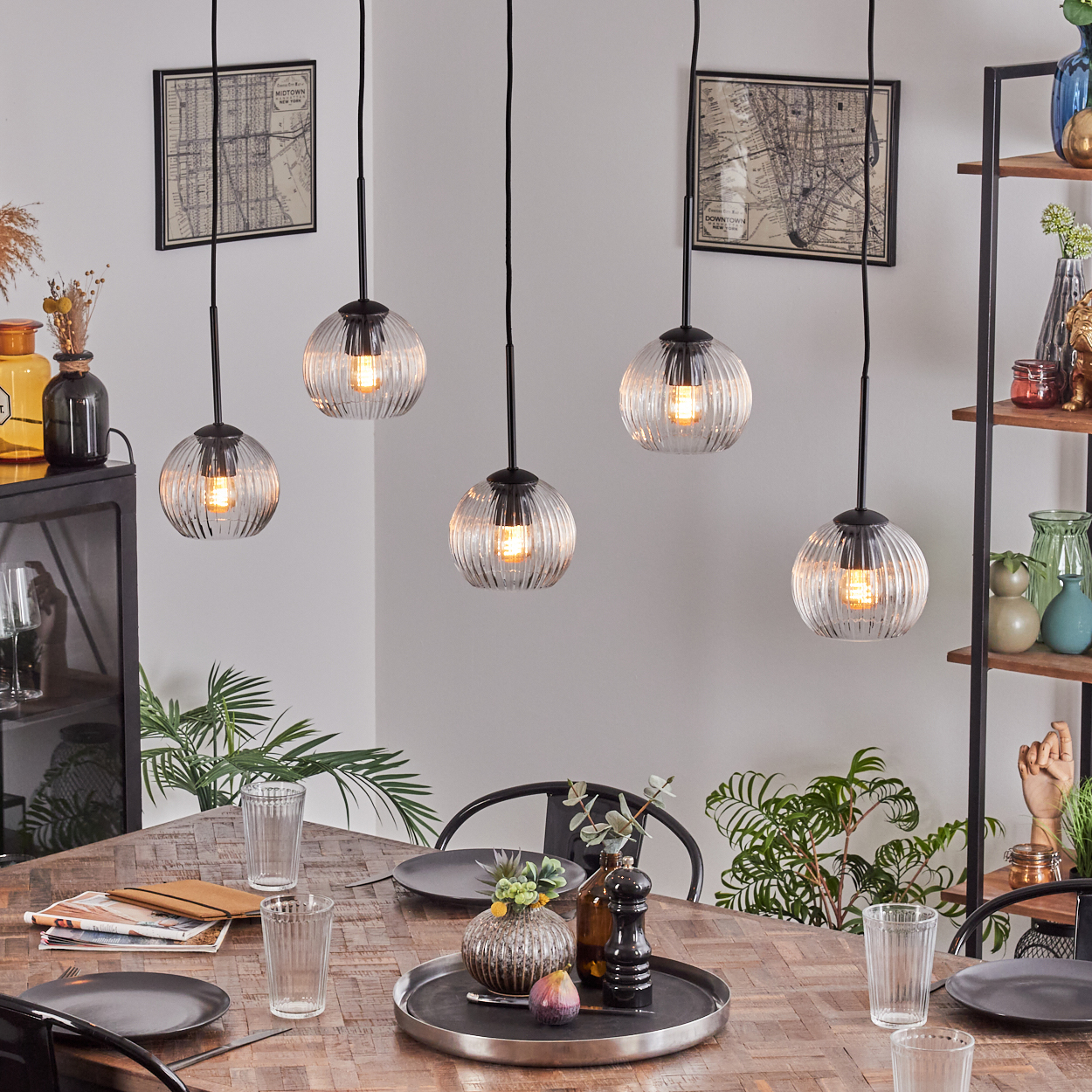Esszimmer mit Holztisch, Glaslampen und Gedecken. Pflanzen und Regale mit Dekorationsartikeln sind im Hintergrund.