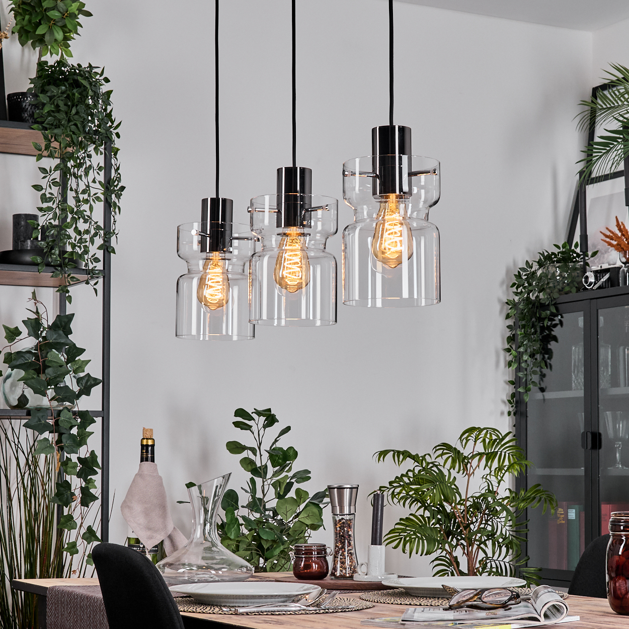 Esszimmer mit Hängelampen. Tisch mit Tellern, Gläsern und Dekorationen. Pflanzen und Regale im Hintergrund.