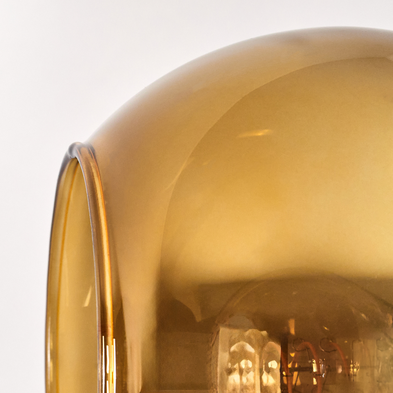 Nahaufnahme eines Lampenschirms aus Glas mit goldenem Farbton. Im Inneren ist eine Glühbirne teilweise sichtbar. Der Hintergrund ist schlicht weiß.