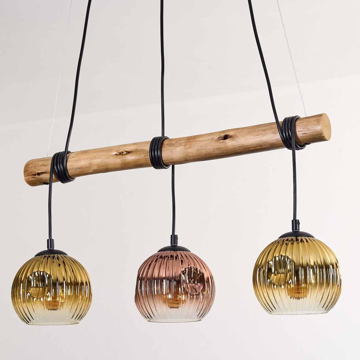 Drei Pendelleuchten mit Glaskugeln hängen an einem Holzbalken. Die Kugeln sind gold- und roségoldfarben. Drähte und eine weiße Wand sind sichtbar.