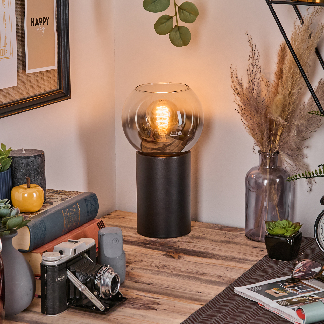 Ein Schreibtisch mit Lampe, Büchern, Kamera und Dekorationen. Die Lampe hat einen Glasschirm. Im Hintergrund eine Vase mit Pflanzen.