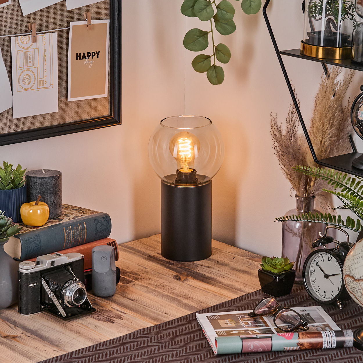 Ein Schreibtisch mit Lampe, Büchern, Kamera, Uhr und dekorativen Gegenständen. Eine Pinnwand hängt im Hintergrund.