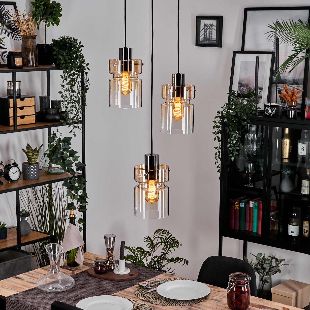 Esszimmer mit drei Hängelampen. Holztisch für das Abendessen gedeckt. Regale mit Pflanzen und Dekor. Schwarzer Schrank.