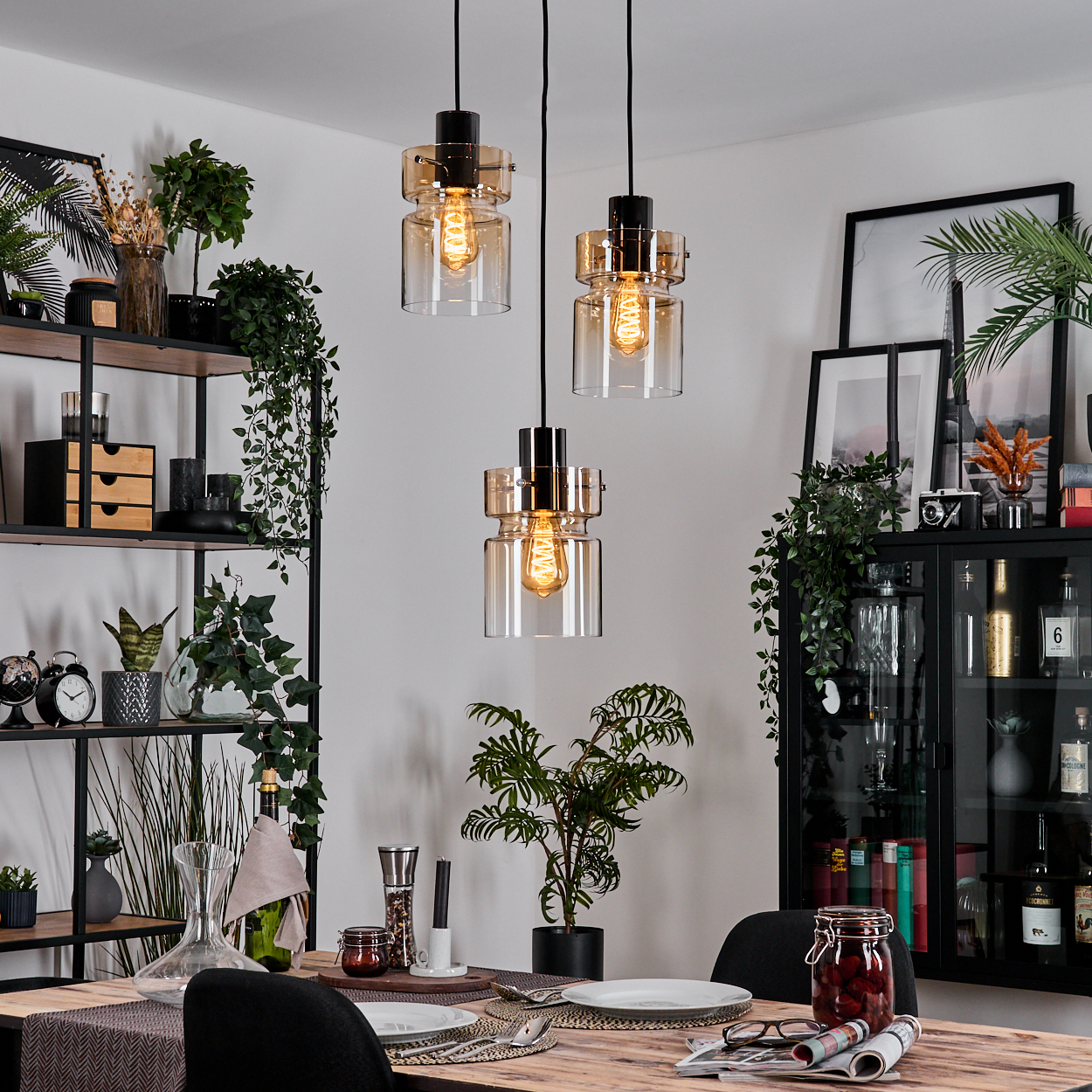 Esszimmer mit Hängelampen. Tisch mit Tellern und Gläsern, schwarze Stühle. Regale und Schrank sind mit Pflanzen sichtbar.