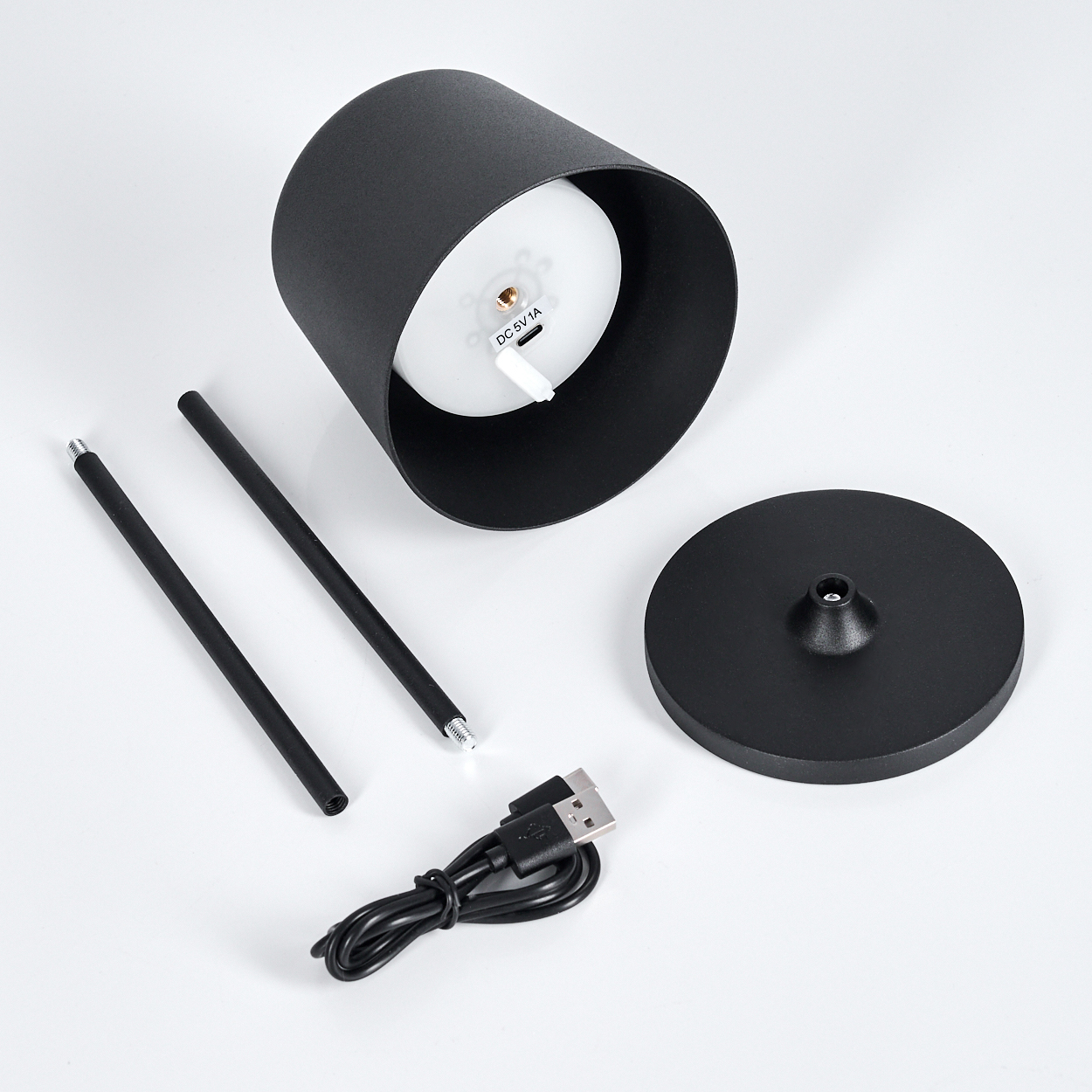 Eine zerlegte schwarze Lampe mit Teilen: zylindrischer Körper, Sockel, zwei Stäbe und ein USB-Kabel, alles auf weißem Untergrund.