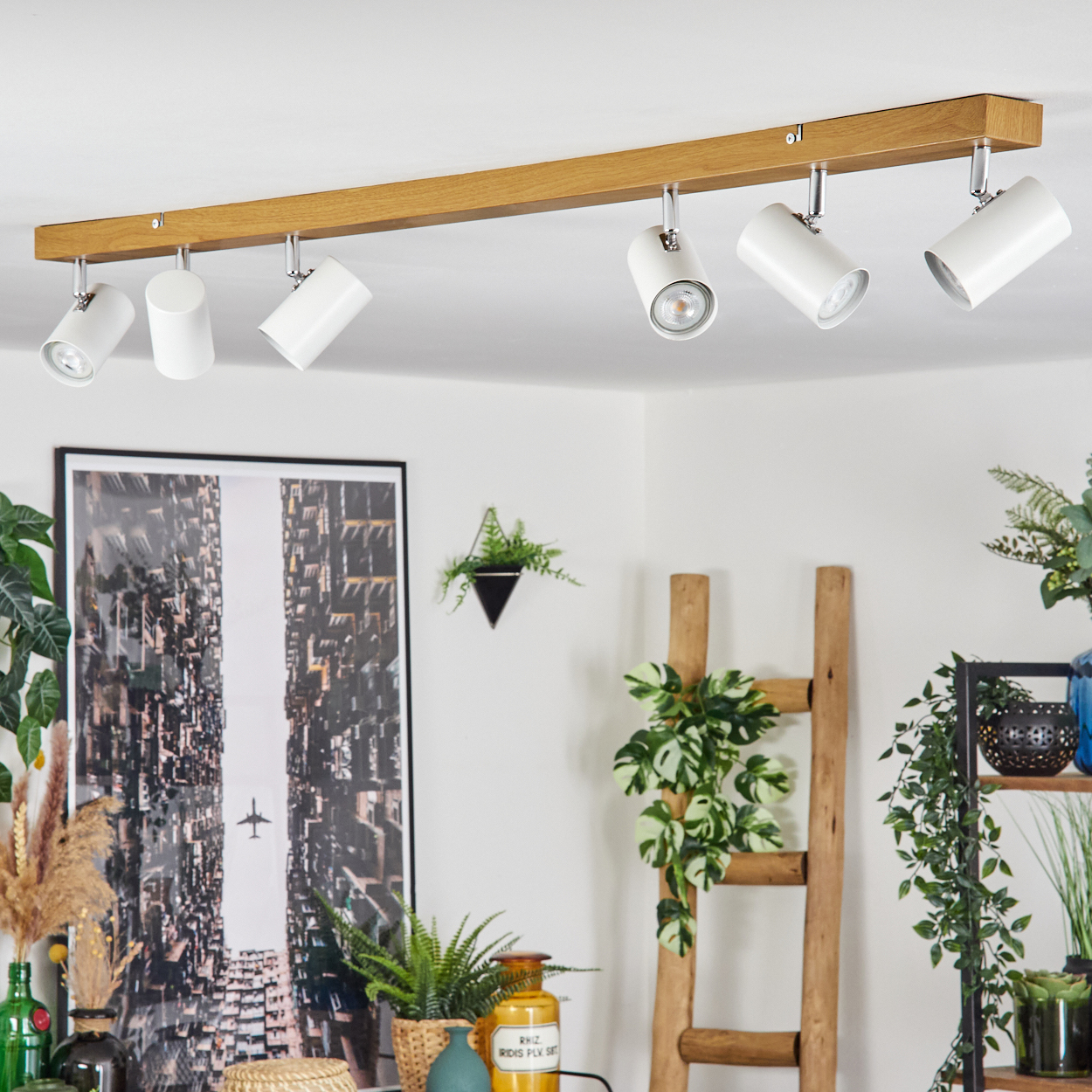 Eine weiße Deckenleuchte mit fünf verstellbaren Strahlern auf einem Holzbalken. Pflanzen, eine Leiter und ein gerahmtes Bild sind im Hintergrund.