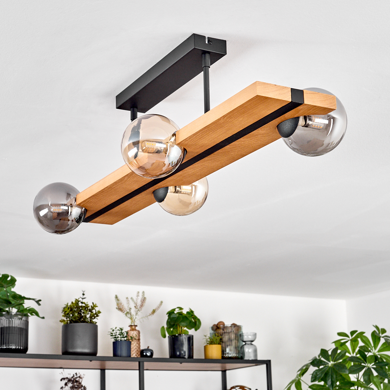 Eine moderne Deckenlampe mit vier Glaskugellampen und einem Holzbalken, schwarzen Akzenten und Pflanzen auf einem Regal.
