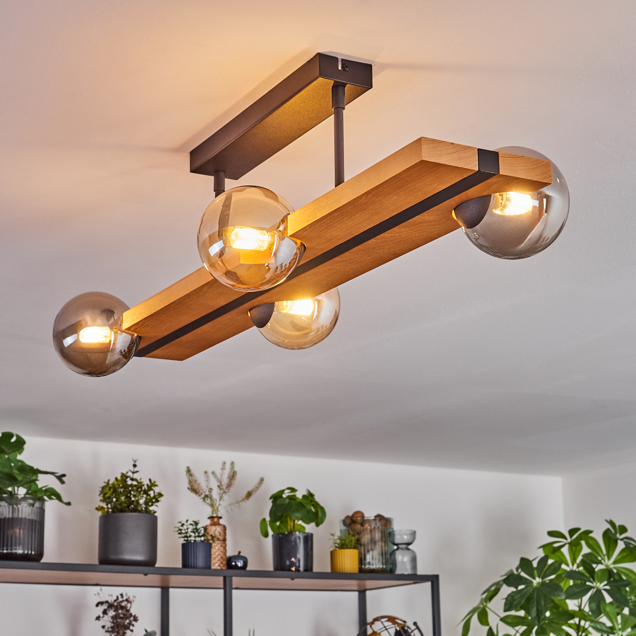 Eine moderne Deckenlampe mit vier Glaskugelleuchten hängt über Topfpflanzen auf einem Regal. Die Lampe hat Holz und dunkles Metall.