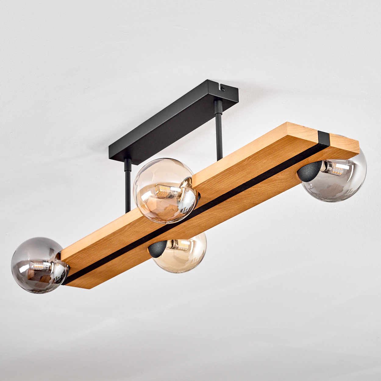 Eine moderne Leuchte mit vier Glaskugeln an einem Holzbalken. Sie hängt an einer schwarzen Metallhalterung an einer weißen Decke.