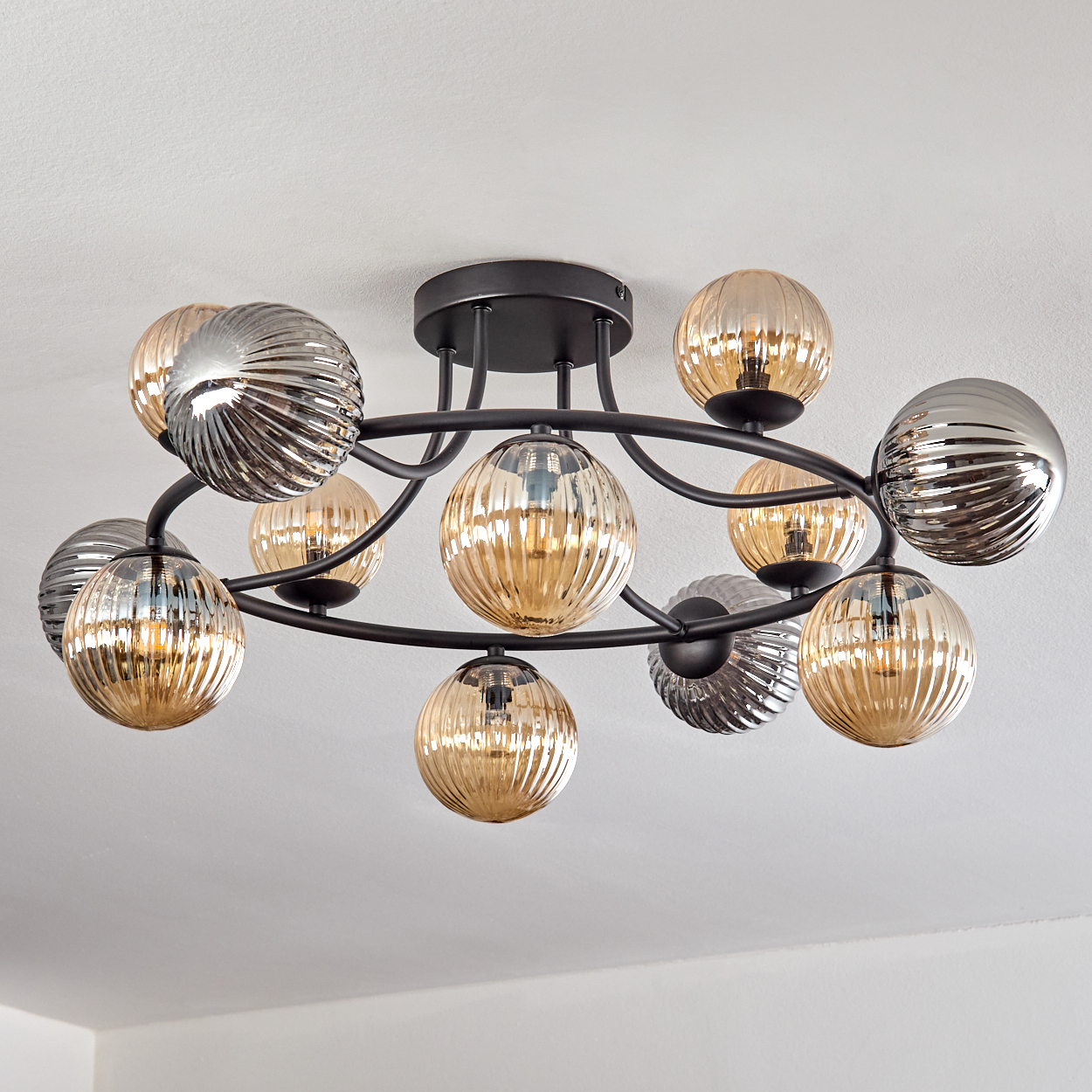 Moderner Kronleuchter mit mehreren kugelförmigen Lichtern. Einige sind bernsteinfarben, andere silberfarben, angeordnet auf einem schwarzen Metallrahmen, gegen eine weiße Decke.