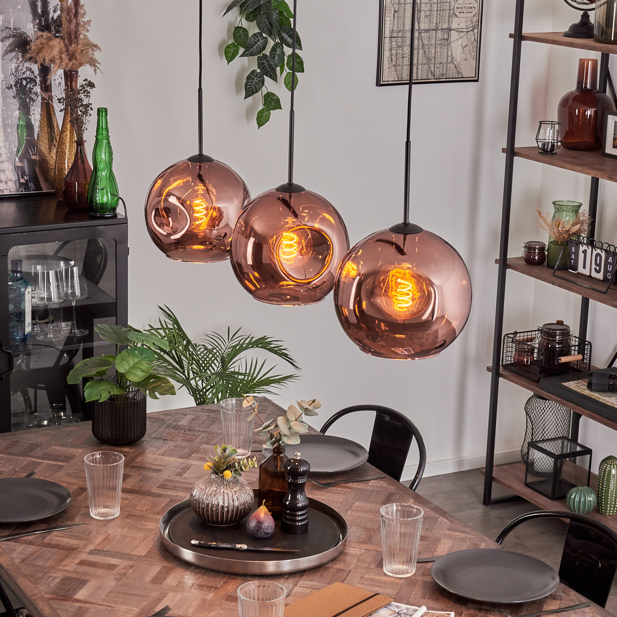 Esszimmer mit Kupfer-Pendelleuchten, gedecktem Tisch und Regalen mit Dekoration. Enthält Pflanzen und einen schwarzen Schrank.