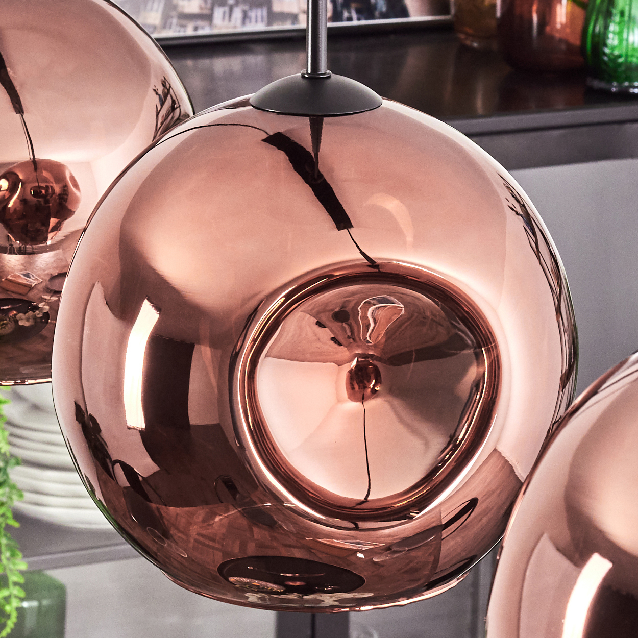 Drei kupferfarbene, kugelförmige Pendelleuchten. Die Lichter spiegeln die umgebenden Objekte wider. Eine schwarze Metallstange hängt jede Leuchte auf.