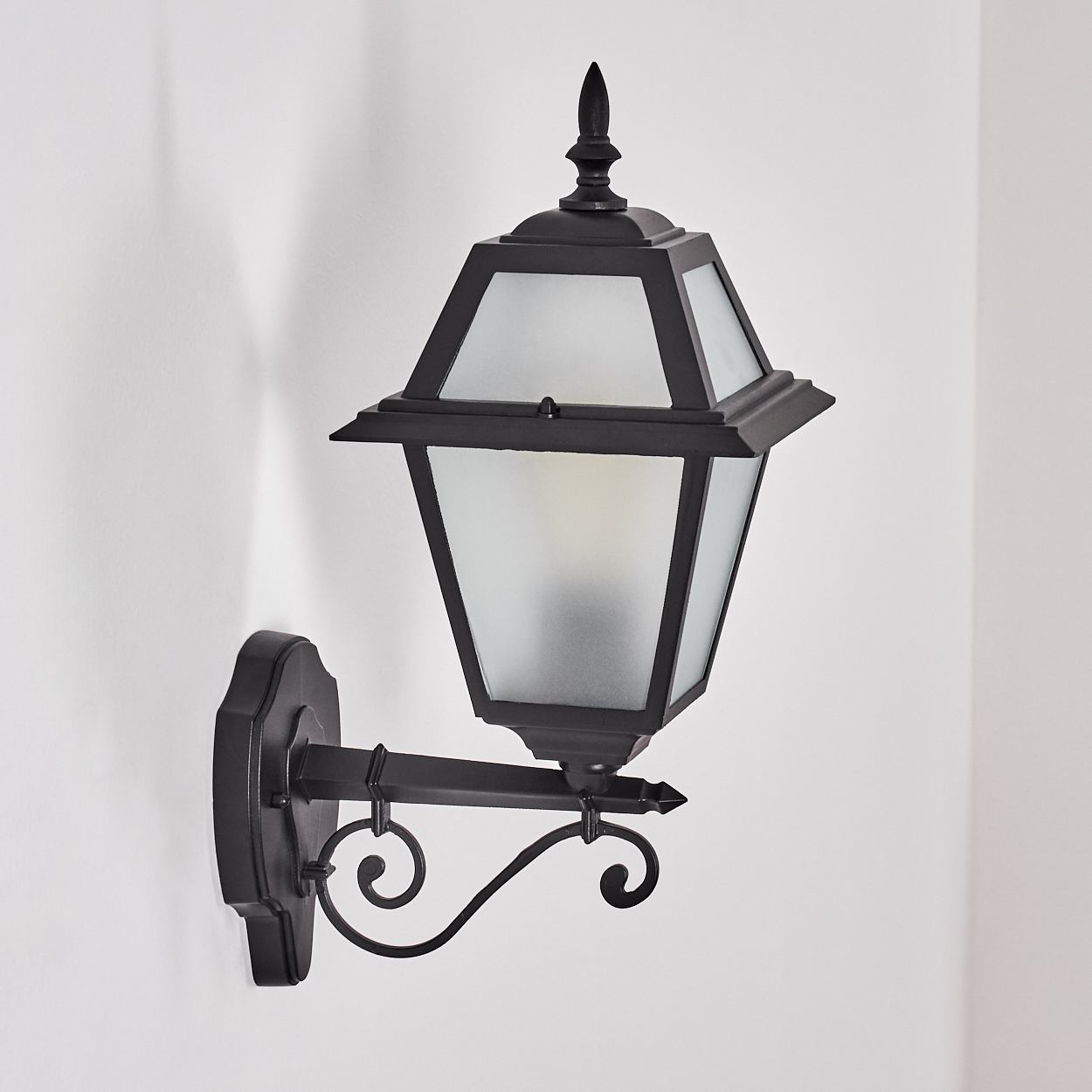 Eine schwarze Außenwandleuchte mit mattiertem Glasschirm ist an einer weißen Wand befestigt. Es hat ein verziertes Design und eine spitze Spitze.