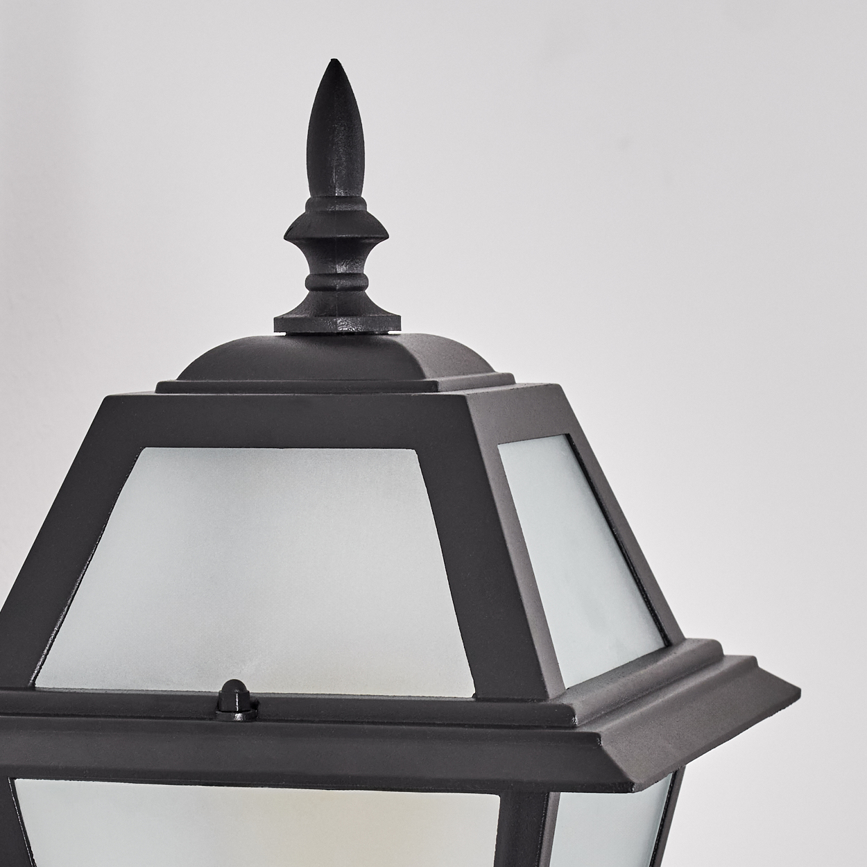 Nahaufnahme einer schwarzen Außenlampe mit Milchglasscheiben. Eine dekorative Spitze krönt die Lampe vor weißem Hintergrund.