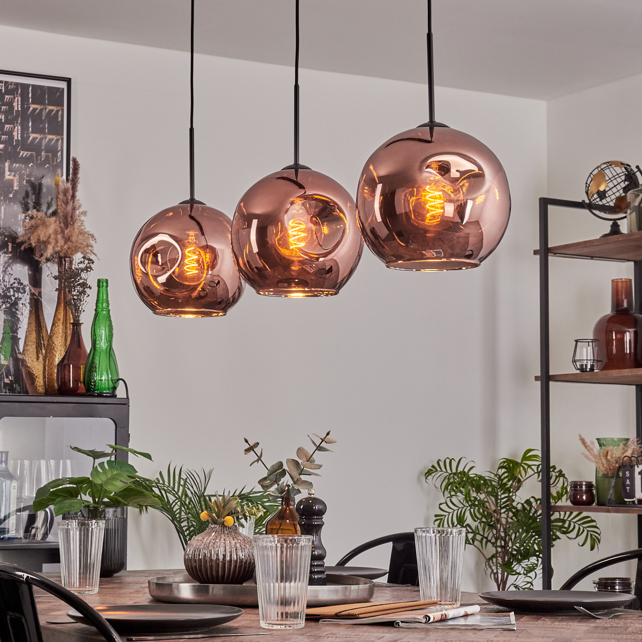 Esszimmer mit kupferfarbenen Pendelleuchten. Tisch mit Tellern und Gläsern. Regale mit Dekorationen. Pflanzen und Flaschen sichtbar.