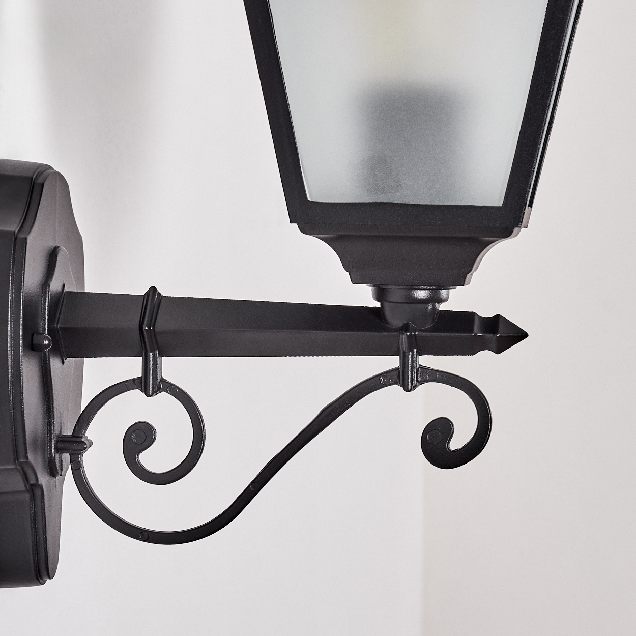 Schwarze Außenlampe. Sie hat einen Laternenstil mit Milchglas und dekorativen Schnörkeln. An einer weißen Wand montiert.