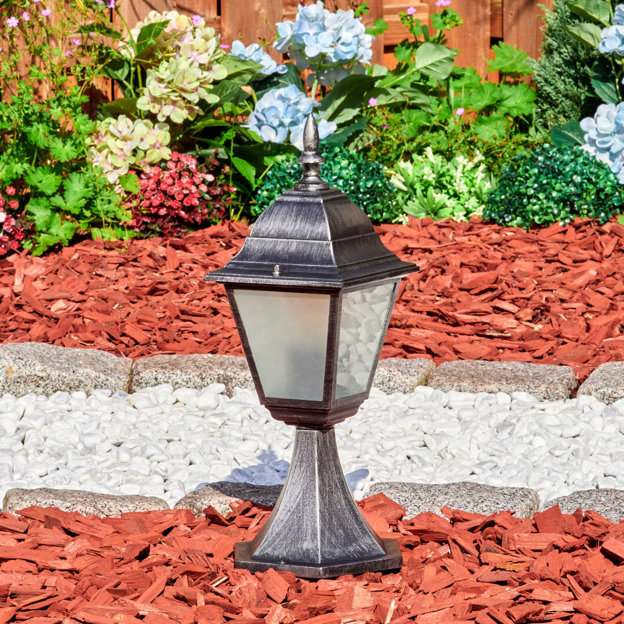 Eine dekorative silberne Lampe steht in einem Gartenbeet mit roter Mulche, weißen Steinen und bunten Blumen im Hintergrund.