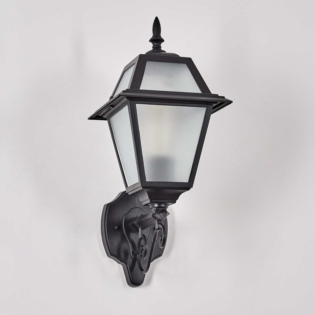 Eine schwarze Außenlampe ist an einer weißen Wand montiert. Die Lampe hat ein Milchglas und eine dekorative Halterung.