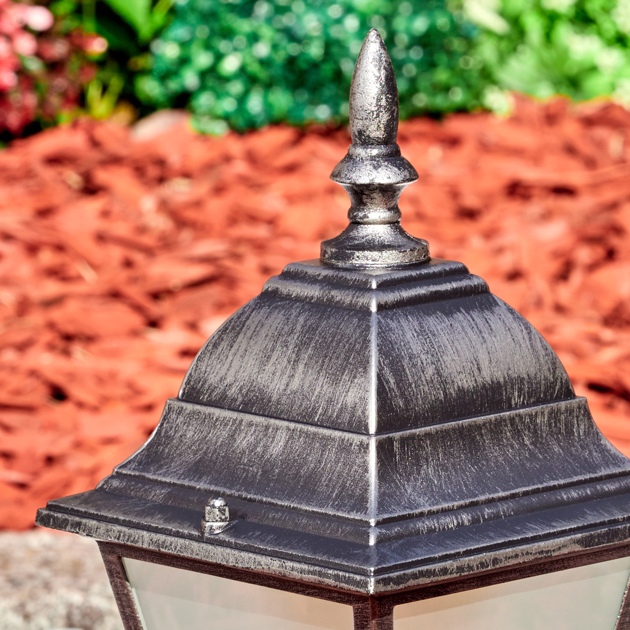 Nahaufnahme einer dekorativen Außenlampe mit silber-bronze-Finish. Rindenmulch und grüne Pflanzen im Hintergrund.