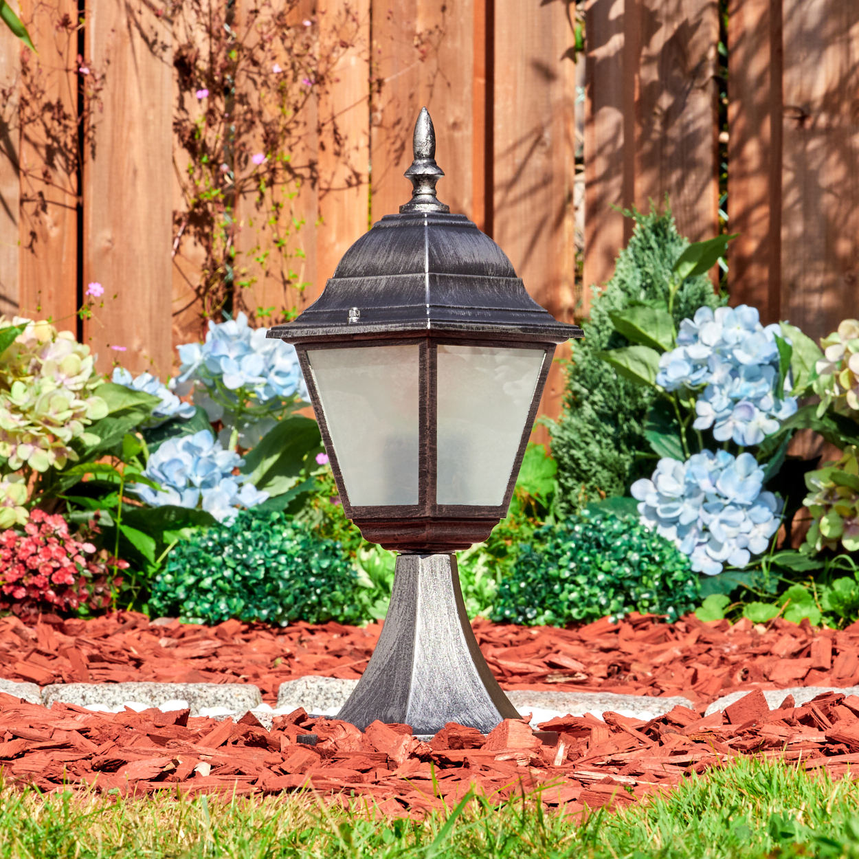 Eine Außenlampe steht in einem Gartenbeet mit roter Mulch, umgeben von Blumen und Grün, mit einem Holzzaun im Hintergrund.