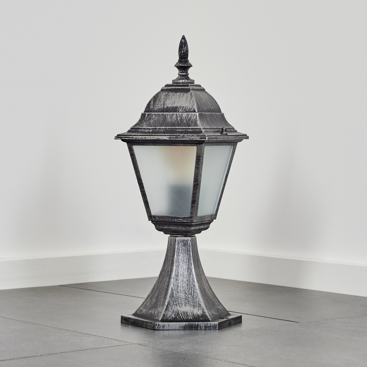 Eine dekorative silberne Außenlampe. Sie hat einen quadratischen Abschnitt aus Milchglas, der auf einem Sockel steht. Der Hintergrund ist weiß.