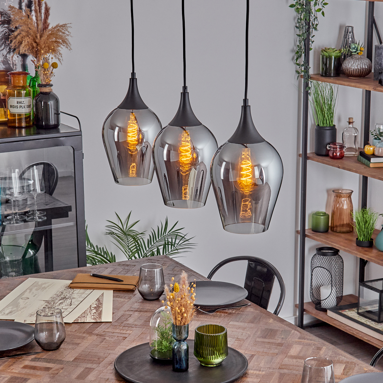 Drei Pendelleuchten hängen über einem Holztisch, der für ein Essen gedeckt ist. Verschiedene Dekorationsartikel und ein Regal im Hintergrund.