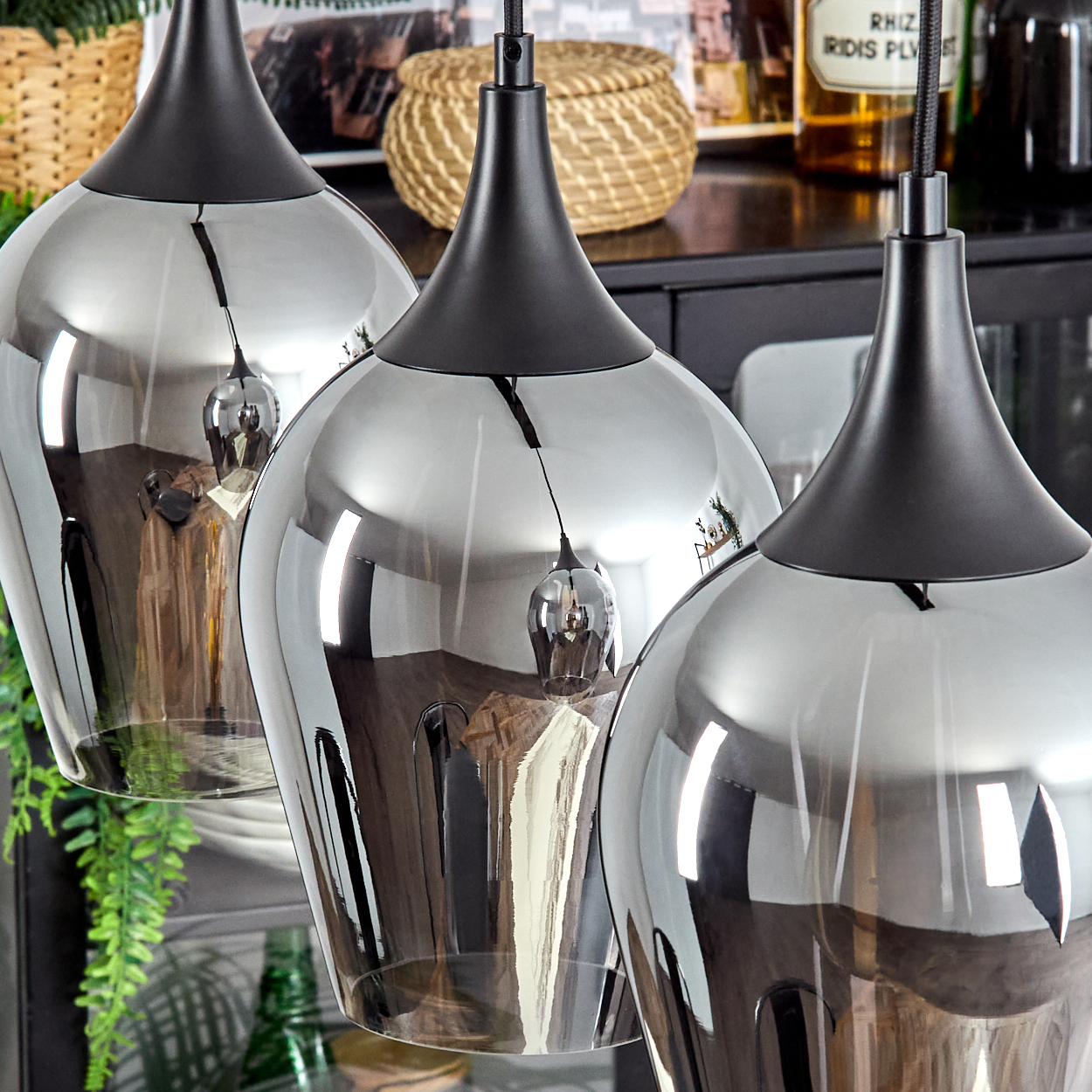 Drei Pendelleuchten mit verspiegelten Glasschirmen hängen. Schwarze Oberteile und Kabel. Hintergrund zeigt Korb, Flaschen und Pflanzen.